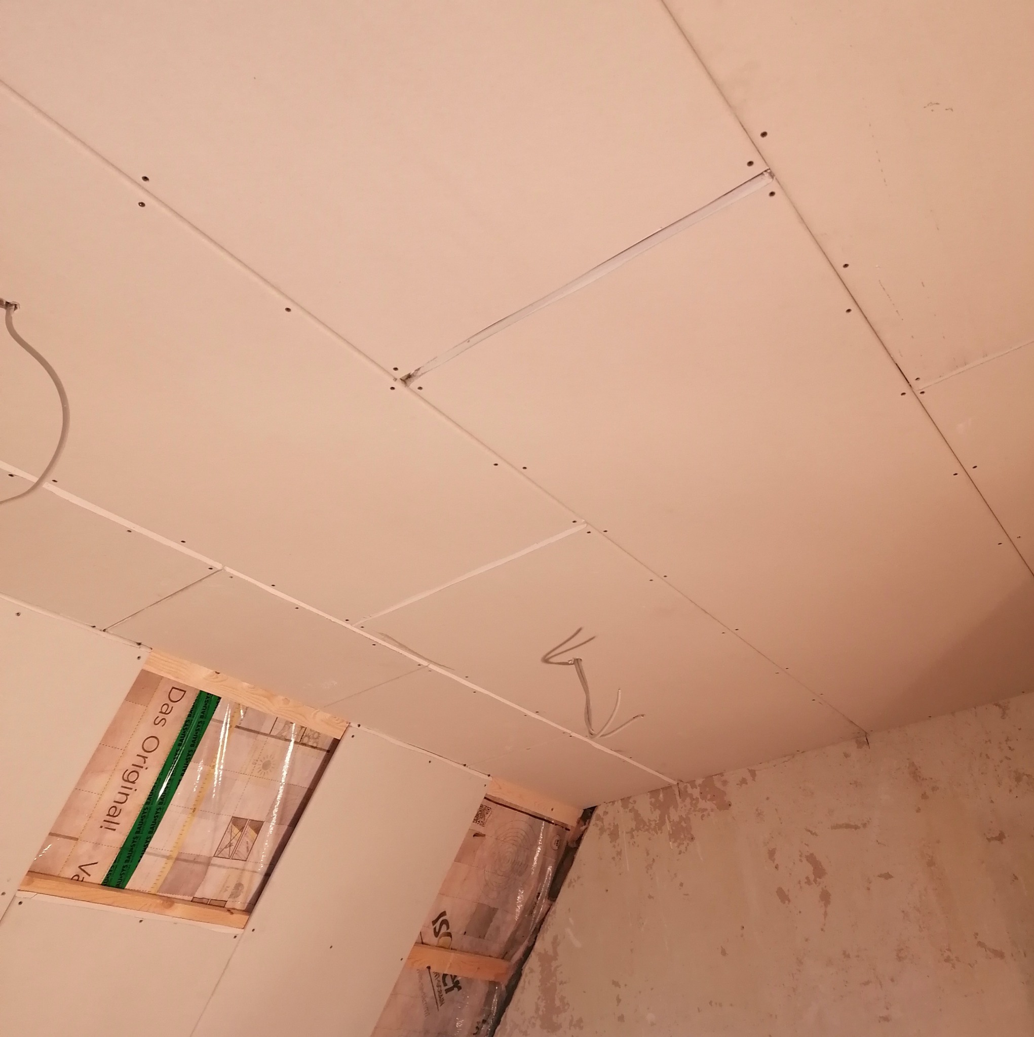 Sufit poddasza z zamontowanymi płytami kartonowo-gipsowymi, widoczne łączenia płyt i wkręty, z fragmentem okna dachowego w trakcie ocieplania wełną mineralną zabezpieczoną folią.