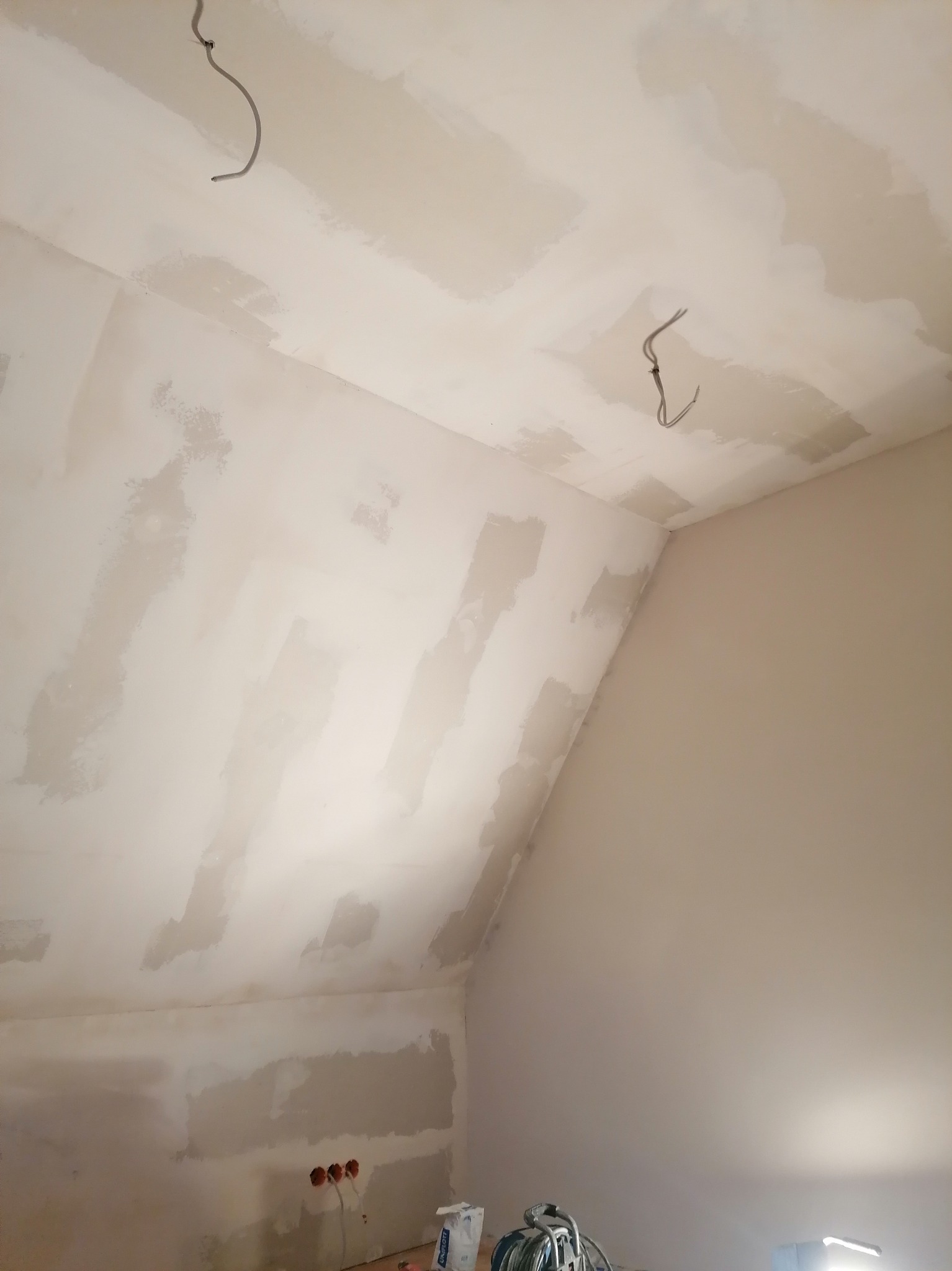 Sufit i ściany poddasza w trakcie szpachlowania, widoczne ślady po łatach gipsowych, wystające przewody elektryczne i puszki instalacyjne, w tle torba z gładzią szpachlową i narzędzia.