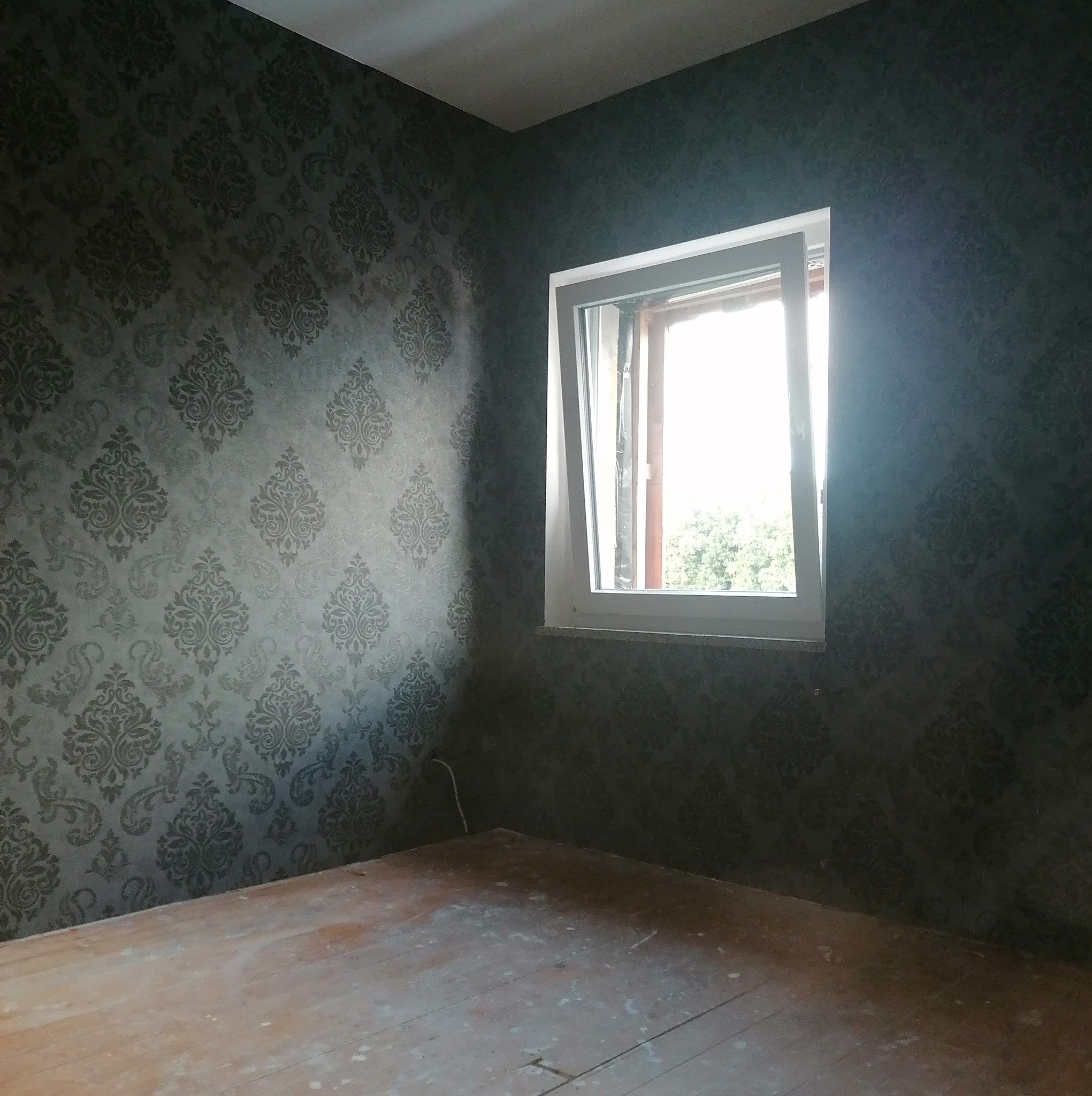 Pokój po malowaniu ścian na ciemny kolor z wzorzystą tapetą, widoczne drewniane podłogi w trakcie renowacji oraz okno z widokiem na zieleń.