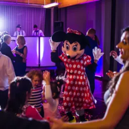 Uśmiechnięta postać w kostiumie Myszki Minnie tańczy z dziećmi i dorosłymi na parkiecie podczas imprezy, w tle DJ i oświetlenie sceniczne.