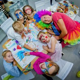 Agencja Opiekunek Cud Niania - Agencja Eventowa - Grupa dzieci z pomalowanymi twarzami rysuje kolorowanki przy stole z animatorem w barwnym stroju i peruce podczas przyjęcia, widok z góry.