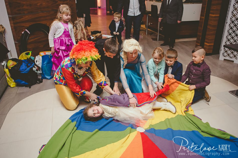 Dziewczynka leżąca na kolorowej chuście trzymanej przez klauna w kolorowym stroju i osobę przebraną za Elsę, obok stoją dzieci w strojach galowych, w tle widoczne osoby dorosłe, sala z elementami...
