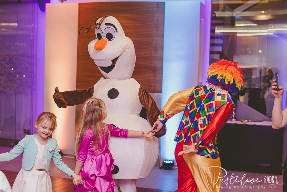 Dwie dziewczynki trzymające się za ręce z osobą w kostiumie Olafa i klauna w kolorowym stroju podczas imprezy.