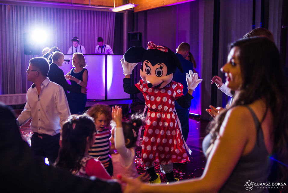 Uśmiechnięta postać w kostiumie Myszki Minnie tańczy z dziećmi i dorosłymi na parkiecie podczas imprezy, w tle DJ i oświetlenie sceniczne.