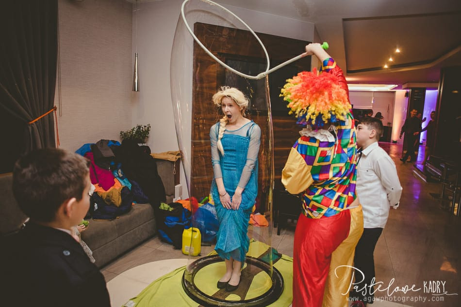 Kobieta przebrana za Elzę z Krainy Lodu wewnątrz dużej bańki mydlanej, obok klaun z kolorową peruką i dziecko w białej koszuli, w tle widoczni inni goście.