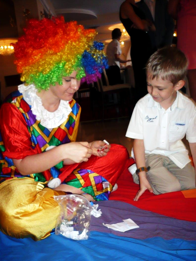 Osoba w stroju klauna z kolorową peruką siedzi na podłodze z dzieckiem, robiąc coś z papieru.
