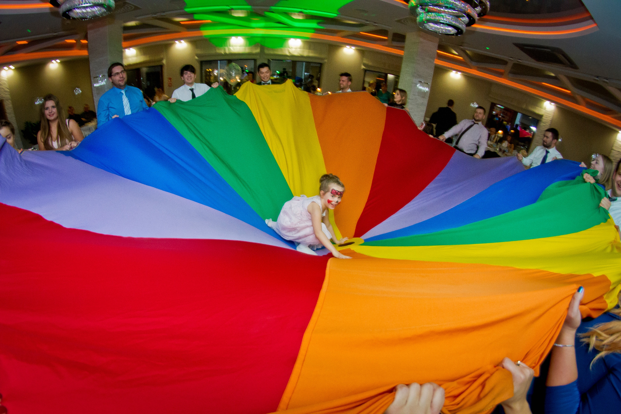 Dziewczynka z pomalowaną twarzą w różowej sukience bawi się na kolorowej chuście trzymanej przez grupę dorosłych w sali bankietowej z dekoracyjnym oświetleniem.