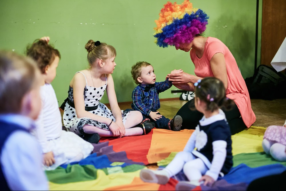 Kobieta w kolorowej peruce gestykuluje do siedzącego na dywanie chłopca podczas zabawy z grupą dzieci w pomieszczeniu z zielonymi ścianami.