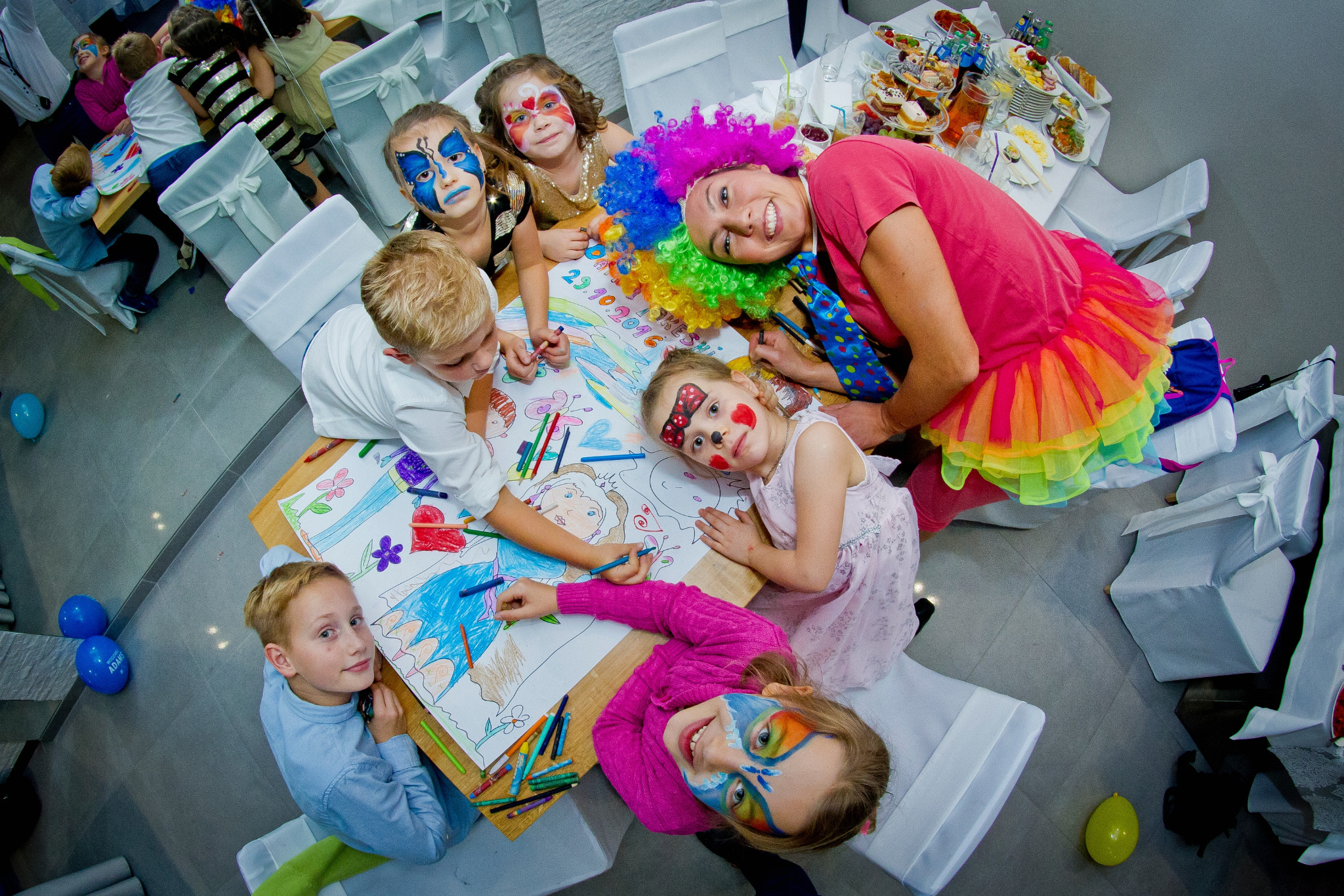 Grupa dzieci z pomalowanymi twarzami rysuje kolorowanki przy stole z animatorem w barwnym stroju i peruce podczas przyjęcia, widok z góry.