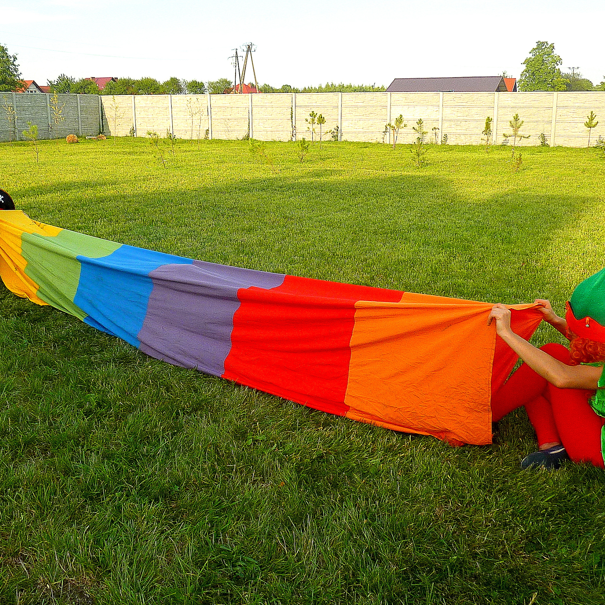 Animator w stroju elfa rozciąga dużą, kolorową chustę animacyjną na trawie podczas zabawy dla dzieci.