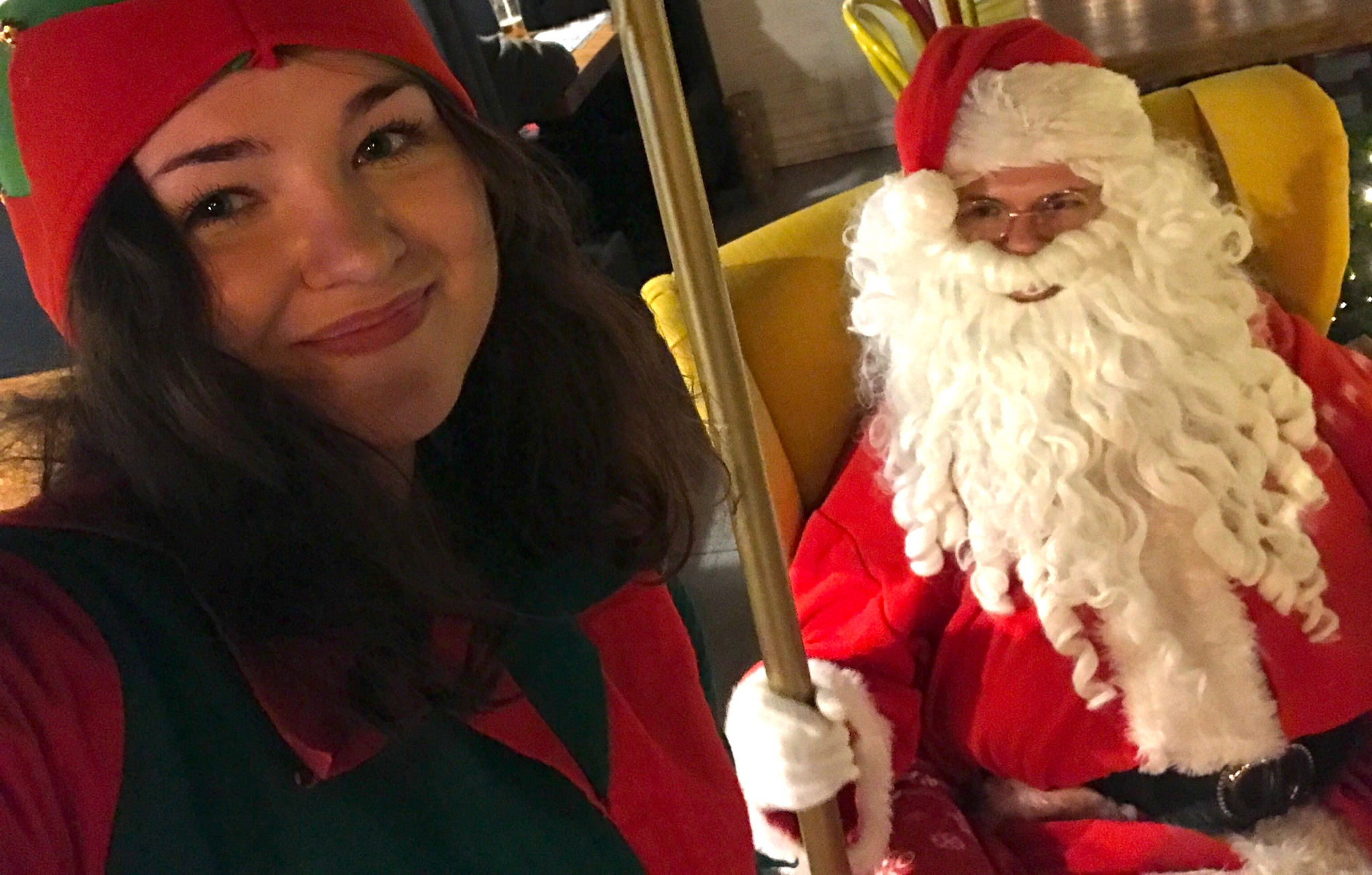 Uśmiechnięta kobieta w stroju elfa i mężczyzna przebrany za Świętego Mikołaja siedzący w żółtym fotelu, trzymający złotą laskę.