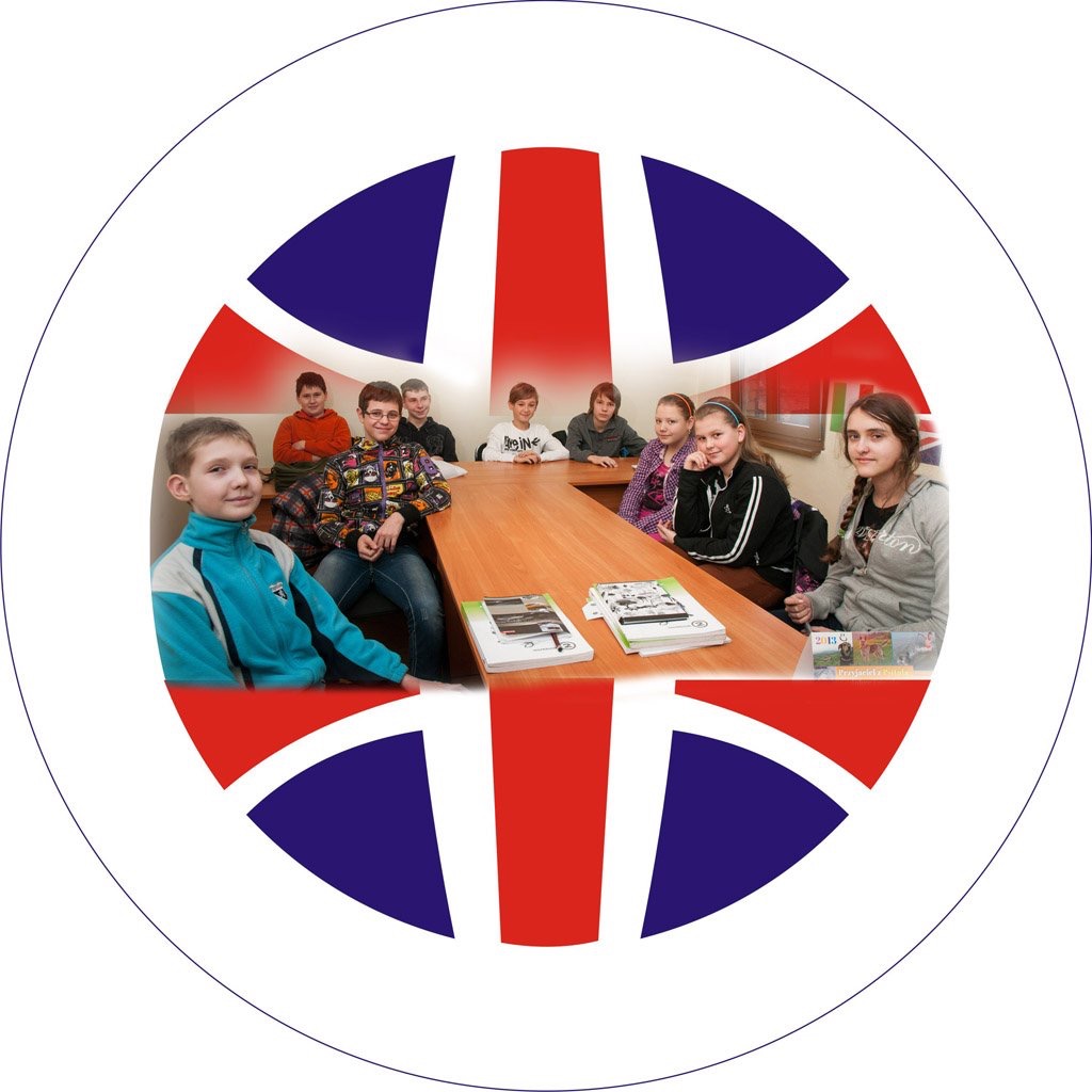 Grupa dzieci siedzi przy drewnianym stole w sali lekcyjnej, na stole leżą podręczniki, w tle flaga Wielkiej Brytanii w okręgu.
