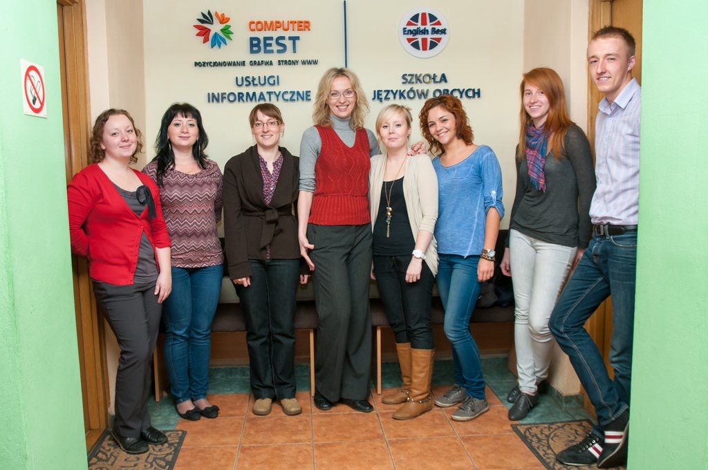Grupa osób, prawdopodobnie pracownicy i uczniowie szkoły językowej, pozuje do zdjęcia na tle logo 'English Best' i 'Computer Best' na ścianie w korytarzu z zielonymi ścianami.