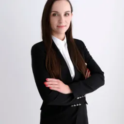 Portret kobiety w czarnym garniturze i białej koszuli, z rękami skrzyżowanymi na piersiach, na jasnym tle.