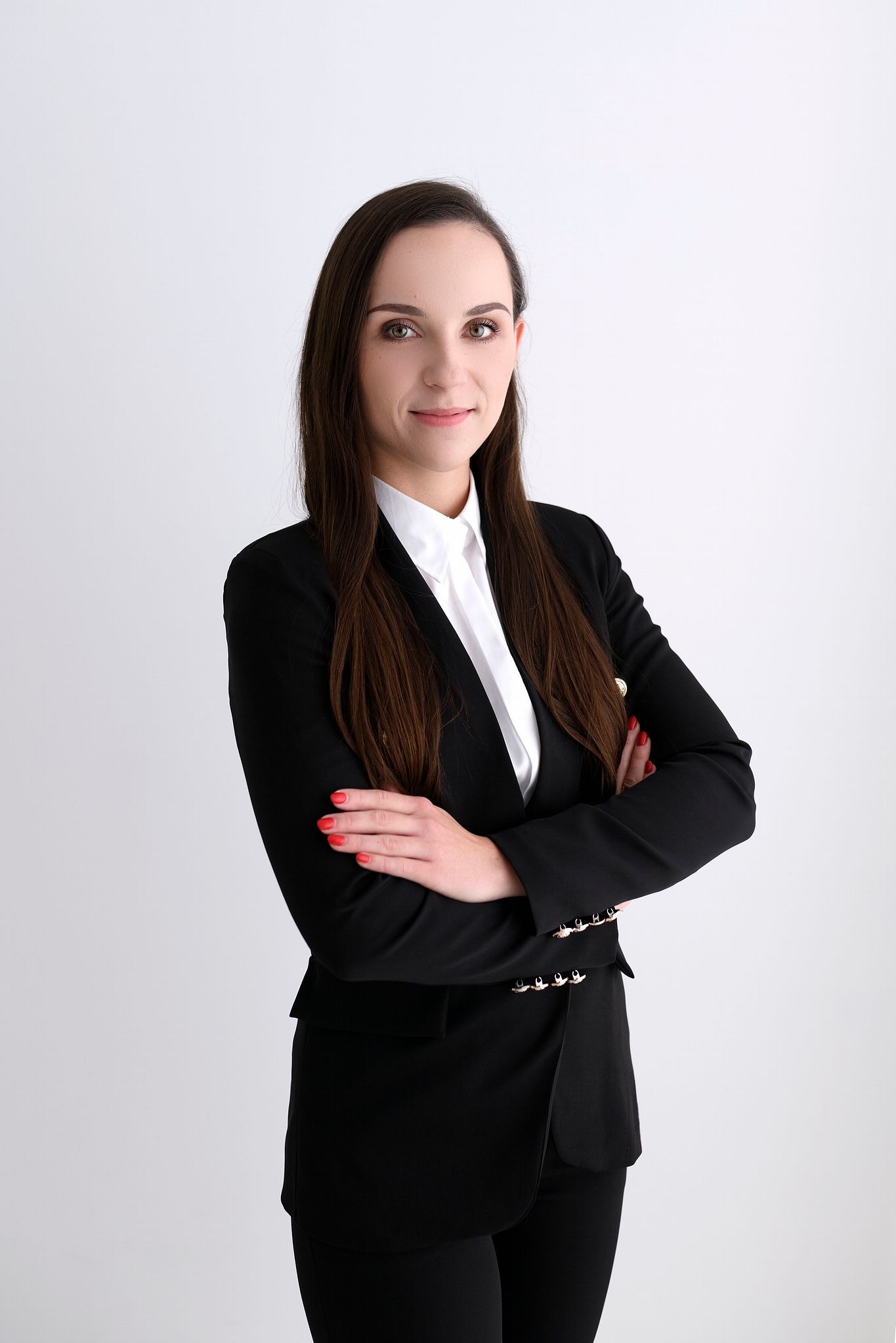Portret kobiety w czarnym garniturze i białej koszuli, z rękami skrzyżowanymi na piersiach, na jasnym tle.