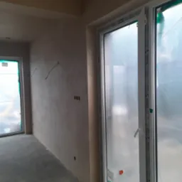 Wnętrze w stanie deweloperskim po nałożeniu gładzi, widoczne wystające przewody elektryczne i okna z folią ochronną.