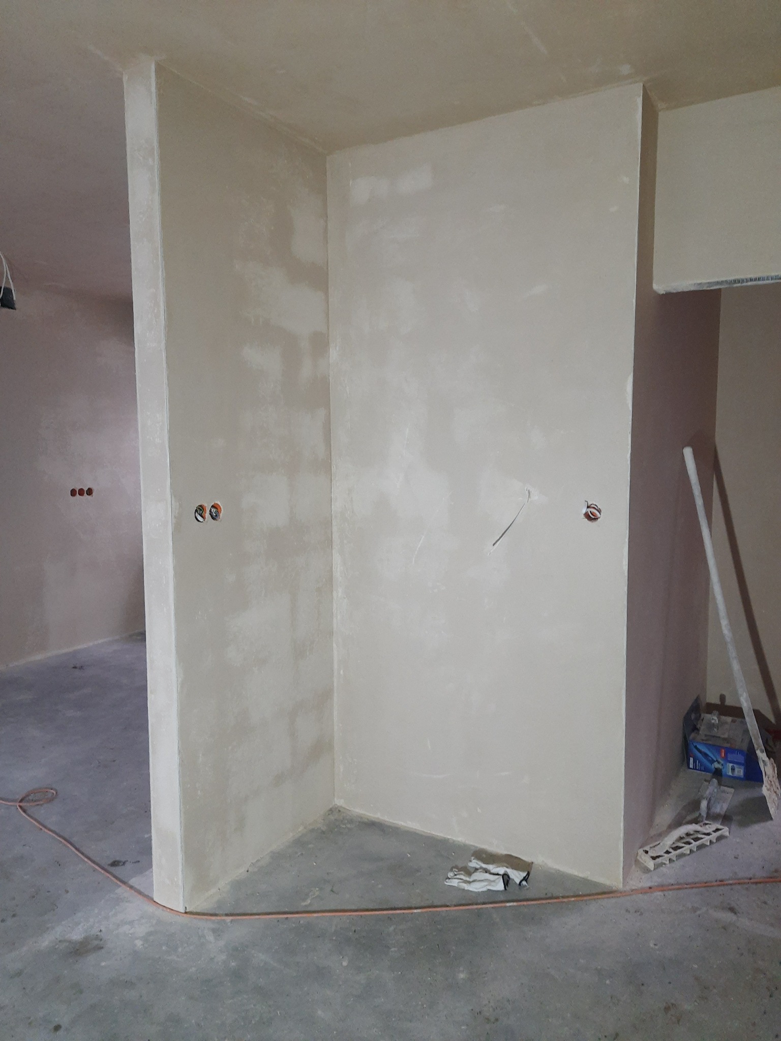 Świeżo nałożony tynk na ścianach wewnętrznych, widoczne puszki instalacyjne, kabel na podłodze i narzędzia w rogu pomieszczenia.