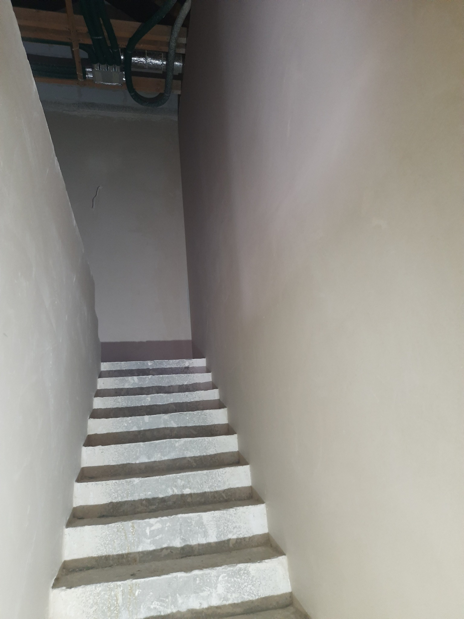 Klatka schodowa w surowym stanie po nałożeniu gładzi, widoczne rury pod sufitem i ściany w jasnym odcieniu szarości.
