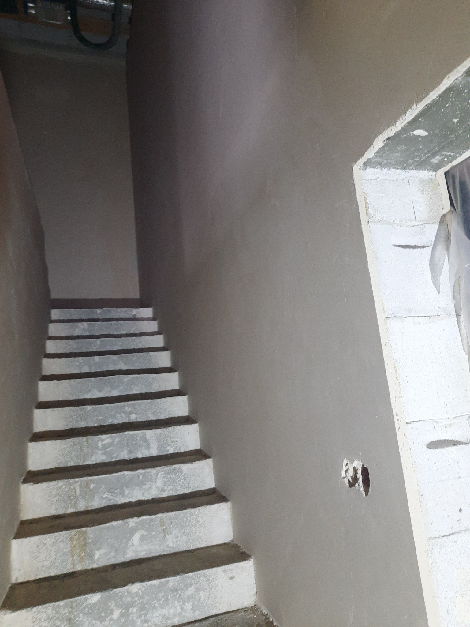 Wnętrze klatki schodowej z surowymi schodami i świeżo nałożonymi, gładkimi tynkami maszynowymi na ścianach, widoczne niedoskonałości i otwór po instalacji elektrycznej.