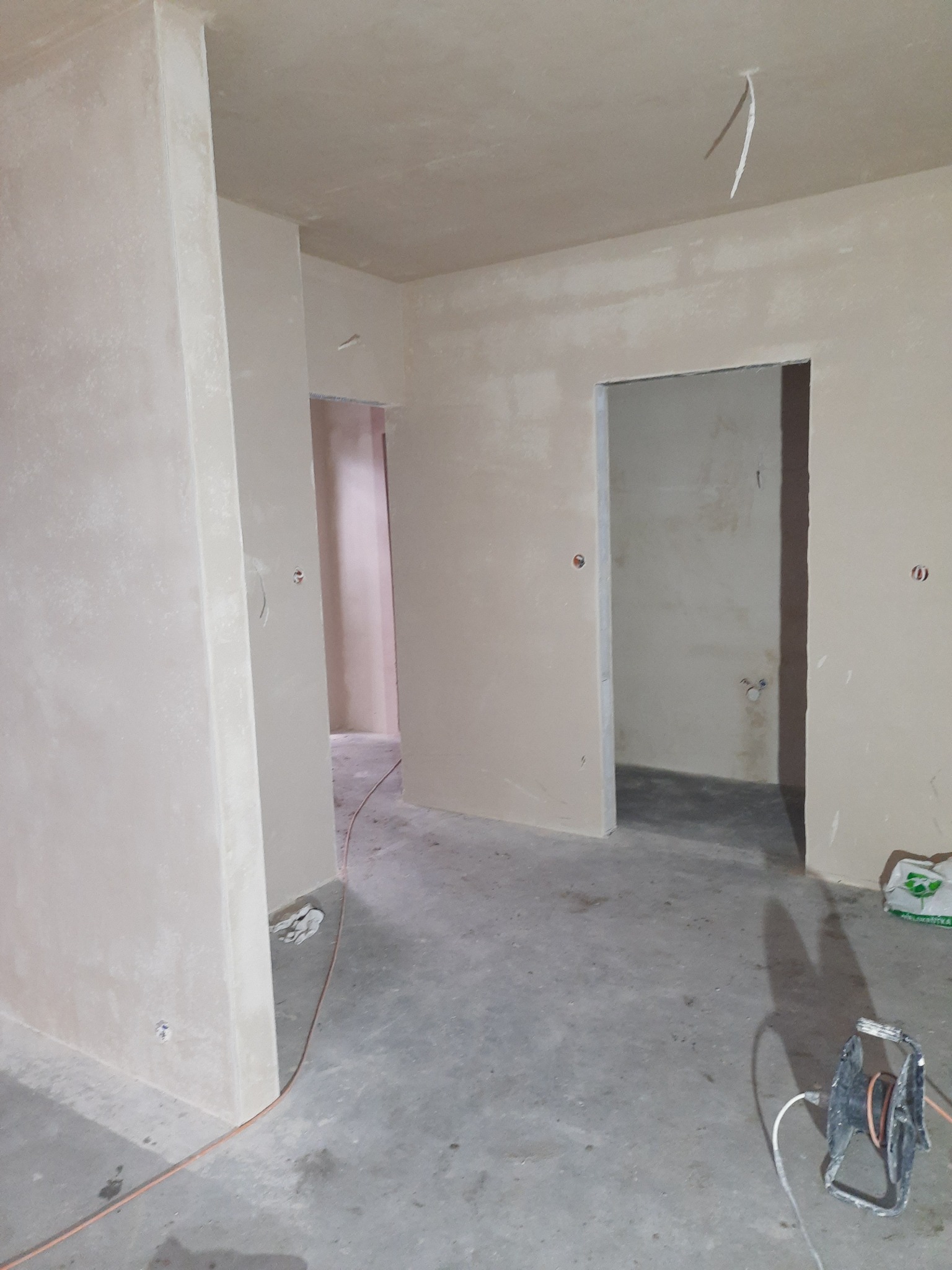 Wnętrze w trakcie prac wykończeniowych, ściany pokryte świeżym, jasnym tynkiem, widoczne przewody elektryczne wystające z sufitu i zwinięty przedłużacz na podłodze.