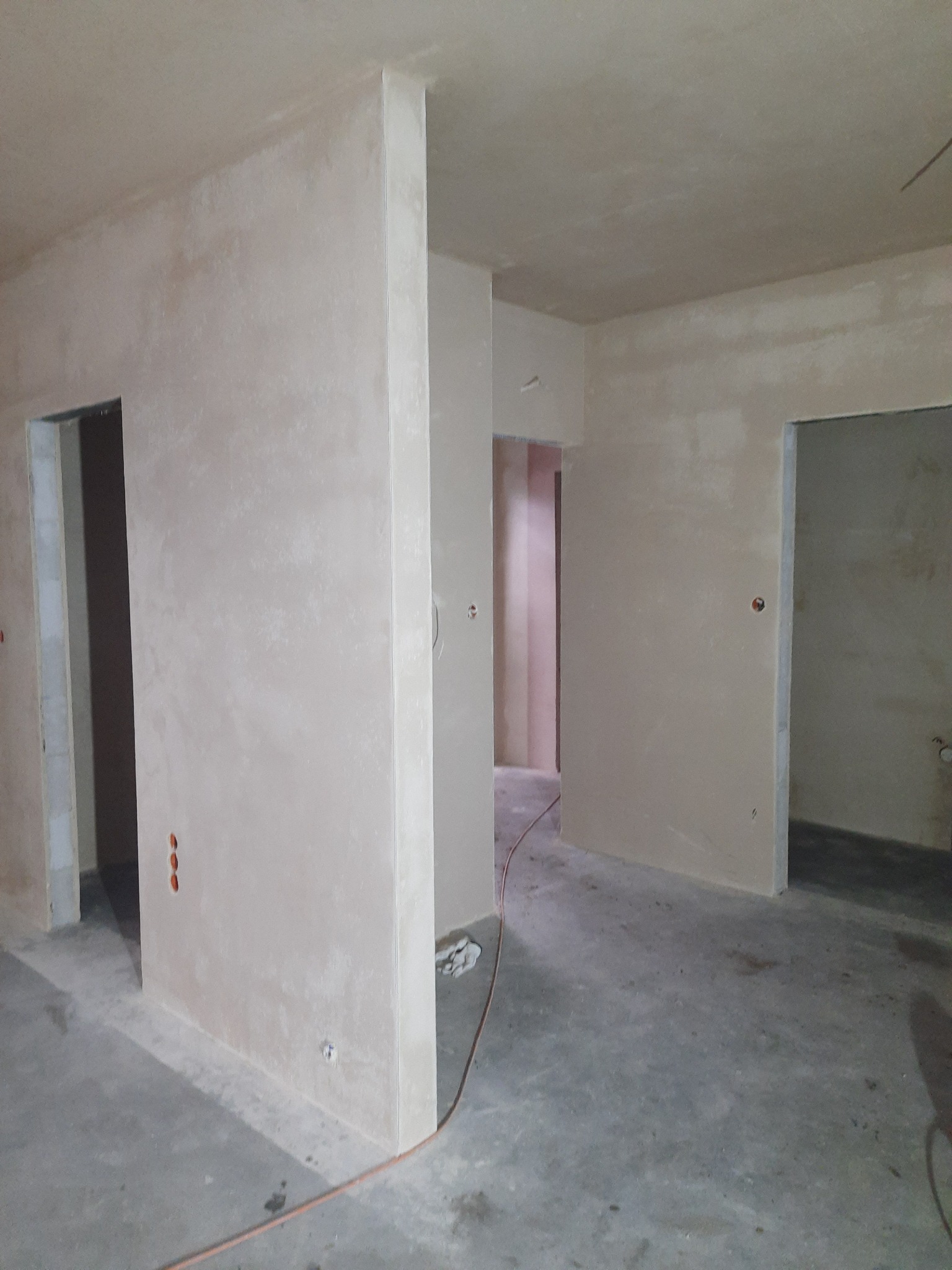 Świeżo położone, jasne tynki maszynowe na ścianach w surowym wnętrzu z widocznymi otworami na gniazdka elektryczne i przewodami na podłodze.