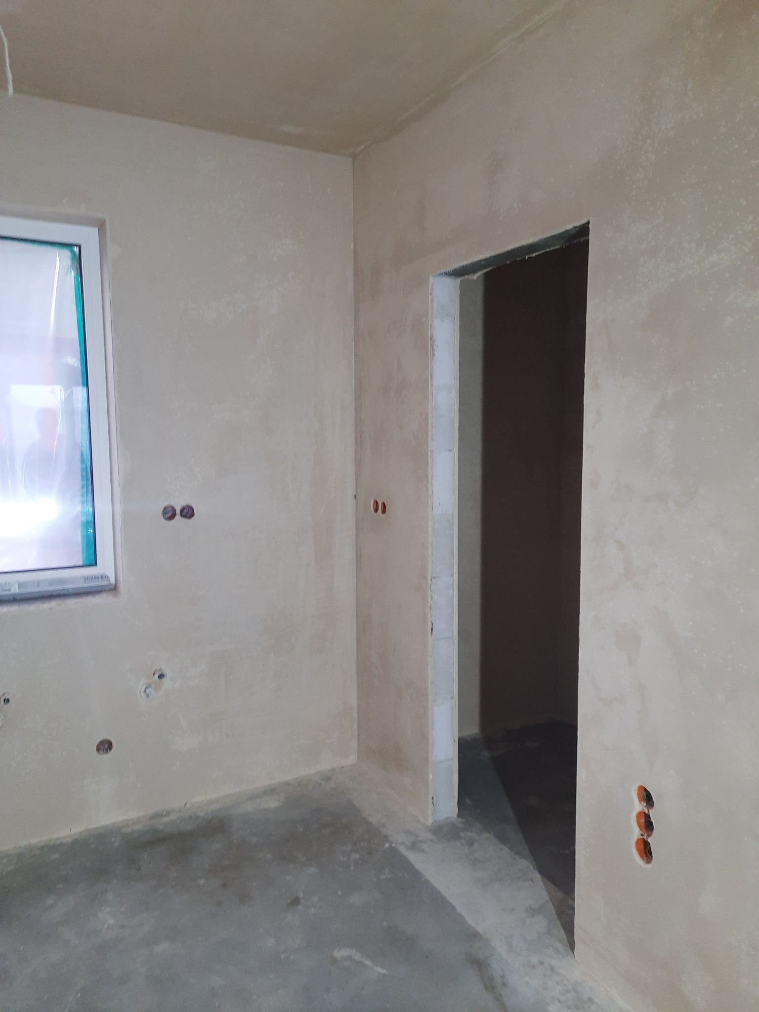 Surowe, jasne wnętrze po nałożeniu tynku, z widocznymi otworami na puszki elektryczne i ościeżnicą drzwiową bez drzwi. Okno zaklejone folią ochronną. Podłoga betonowa.