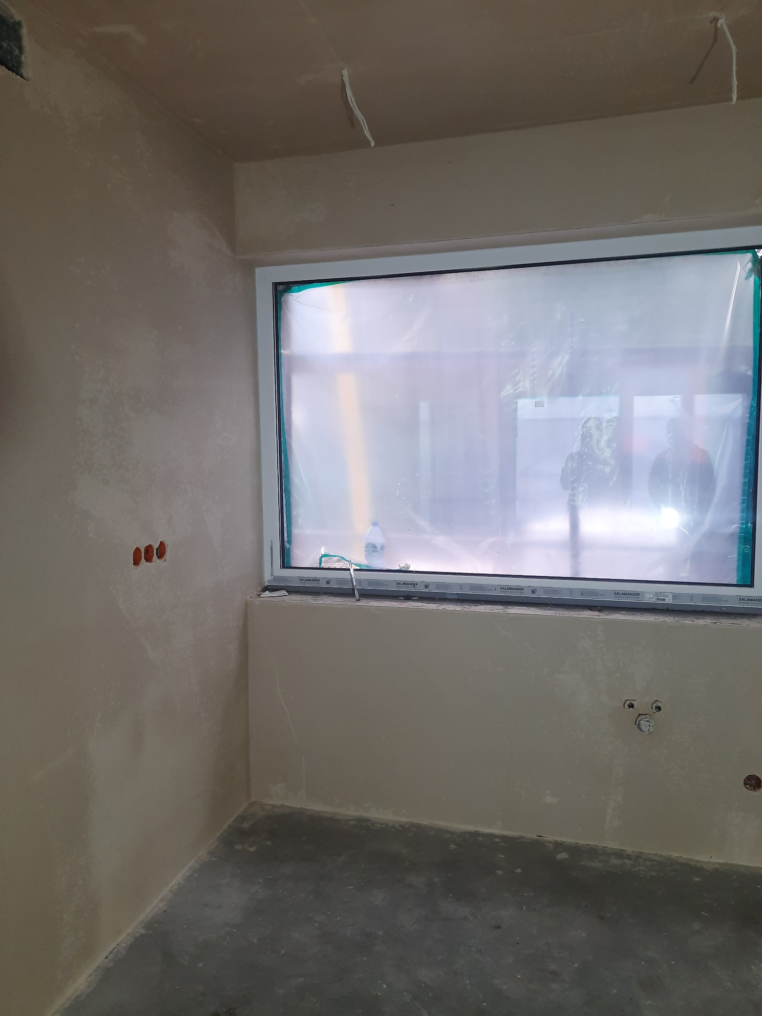 Świeżo otynkowane ściany w pomieszczeniu z oknem zabezpieczonym folią malarską, widoczne puszki elektryczne i instalacja wodna.