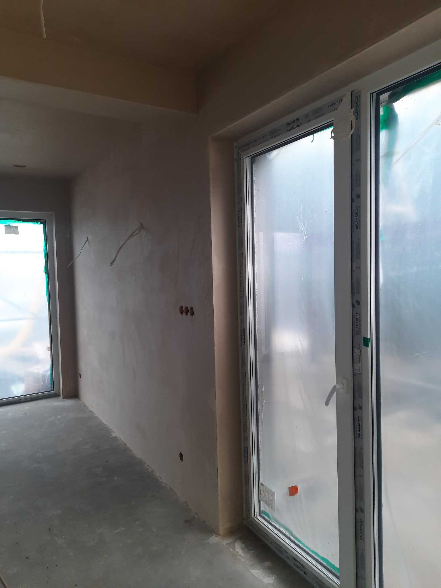 Wnętrze w stanie deweloperskim po nałożeniu gładzi, widoczne wystające przewody elektryczne i okna z folią ochronną.