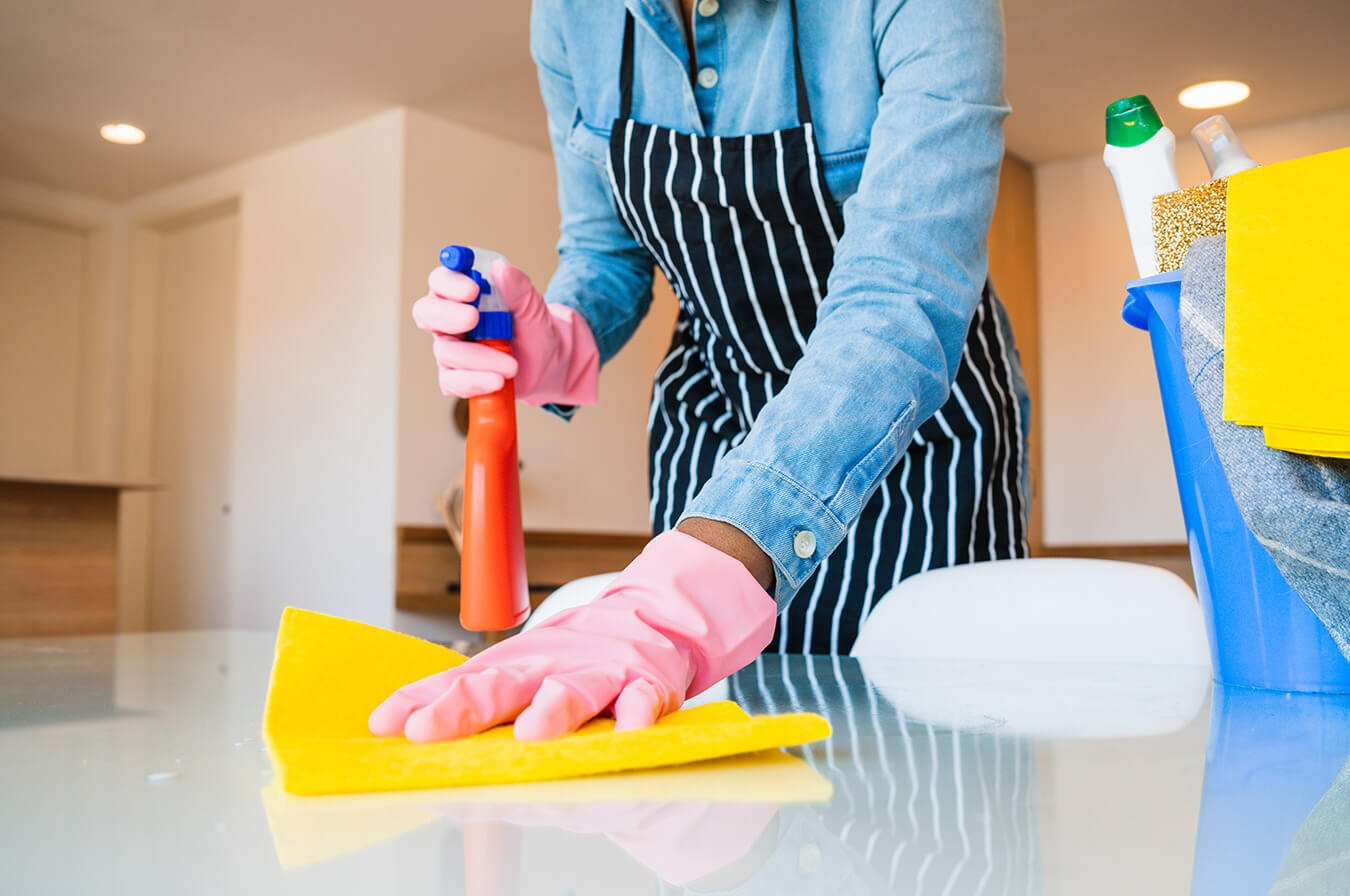 Osoba w różowych rękawiczkach i fartuchu w paski, spryskuje pomarańczowym sprayem i czyści żółtą ściereczką błyszczący biały blat. Obok niebieskie wiadro z detergentami i ściereczkami.