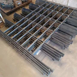 Siatka z metalowych prętów i płaskowników, ułożona warstwami na podłodze, widoczne spawy łączące elementy, w tle paleta z wyciętymi elementami metalowymi.