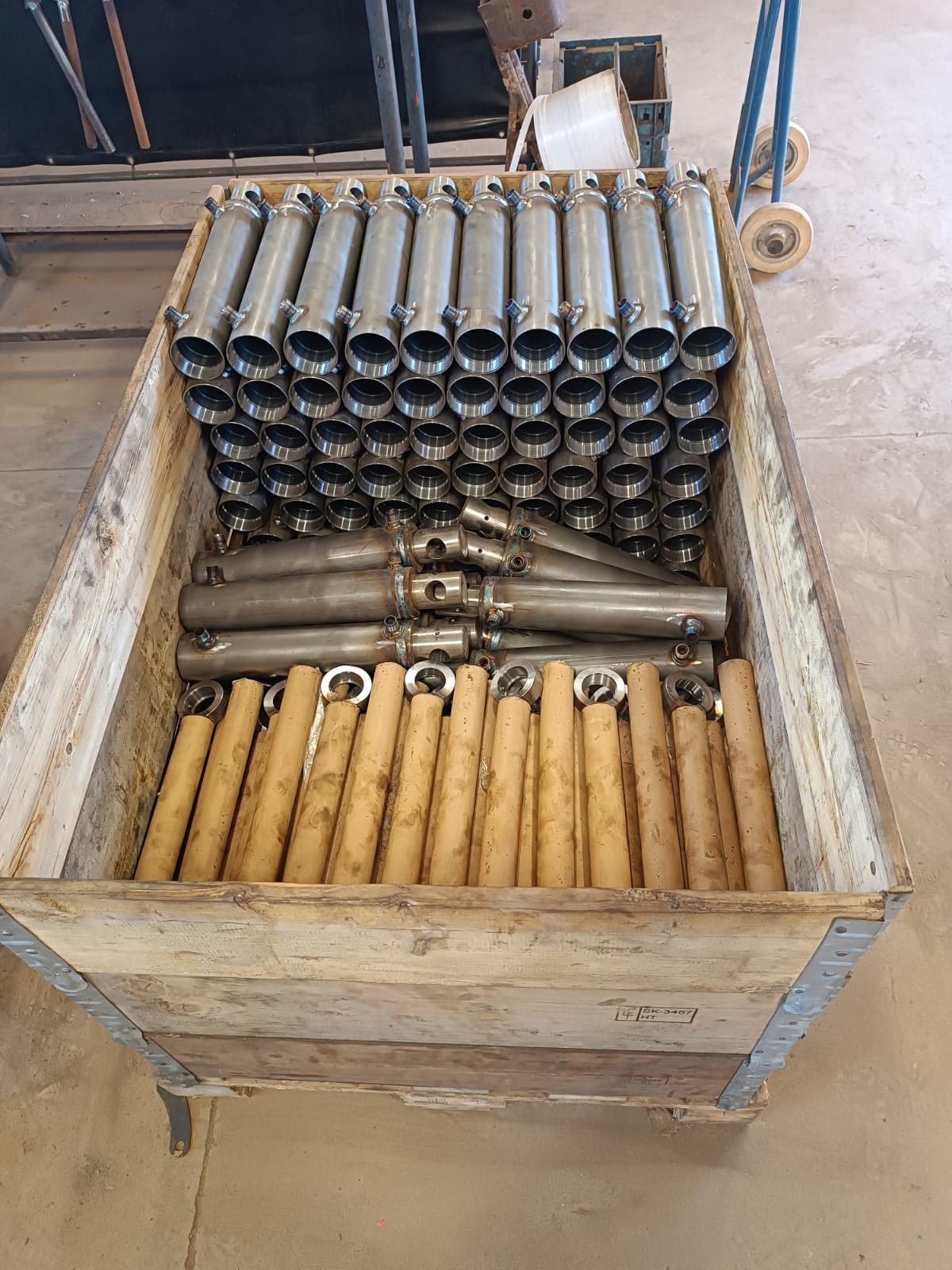 Drewniana skrzynia wypełniona metalowymi cylindrami o różnych wykończeniach: polerowane oraz pokryte ochronną powłoką, przygotowane do dalszej obróbki lub transportu.
