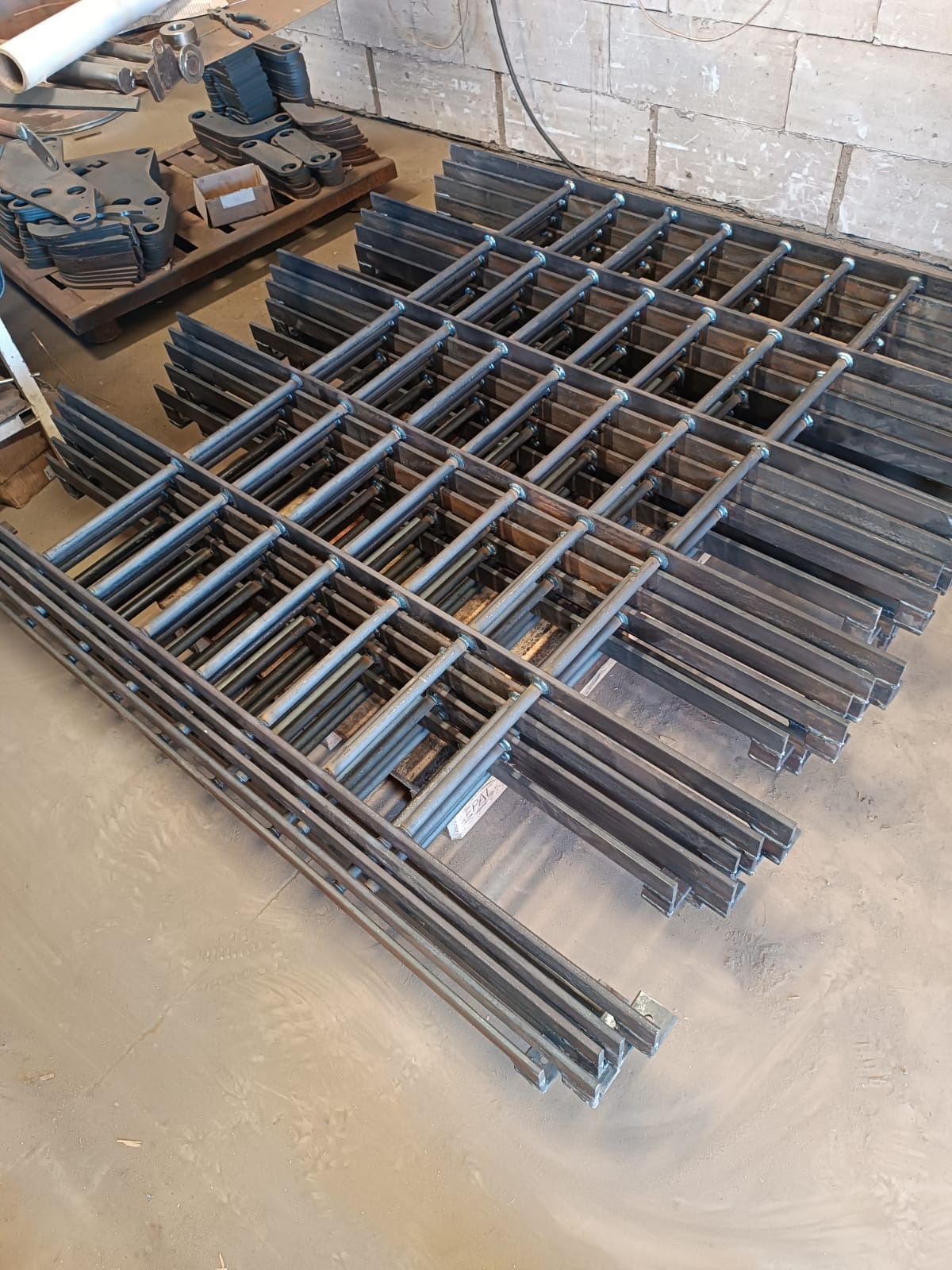 Siatka z metalowych prętów i płaskowników, ułożona warstwami na podłodze, widoczne spawy łączące elementy, w tle paleta z wyciętymi elementami metalowymi.