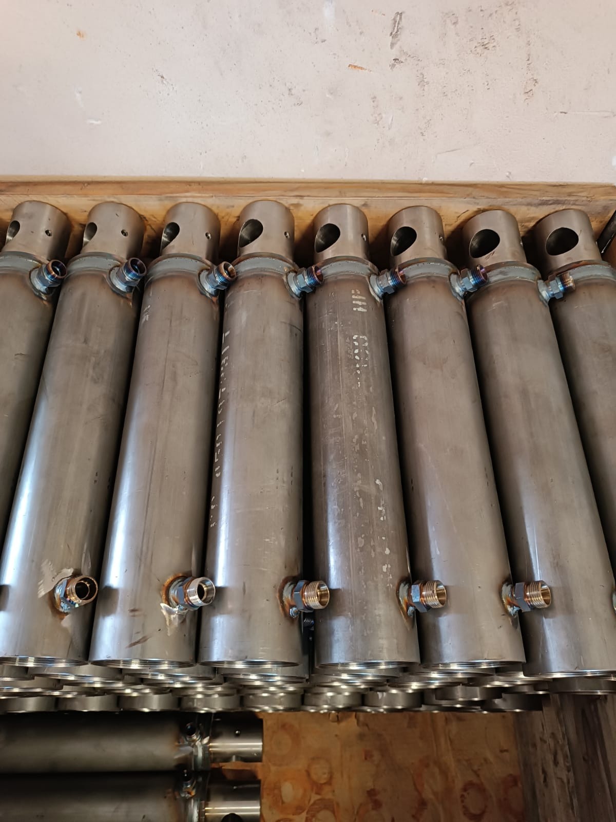 Rząd cylindrycznych, metalowych zbiorników z przyspawanymi złączkami, ułożonych jeden za drugim w drewnianej skrzyni, widoczne ślady po spawaniu.