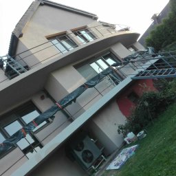 Remonty i wykończenia Oliwier Gacmenga - Dwupiętrowy dom z balkonami w trakcie prac elewacyjnych, widoczna drabina zabezpieczona folią, częściowo pomalowana ściana i sprzęt na trawniku.