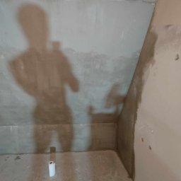 Remonty i wykończenia Oliwier Gacmenga - Ściana pod skosem w trakcie prac wykończeniowych, widoczna siatka na suficie, ślady po gładzi, cień osoby robiącej zdjęcie i rolka papieru na podłodze.