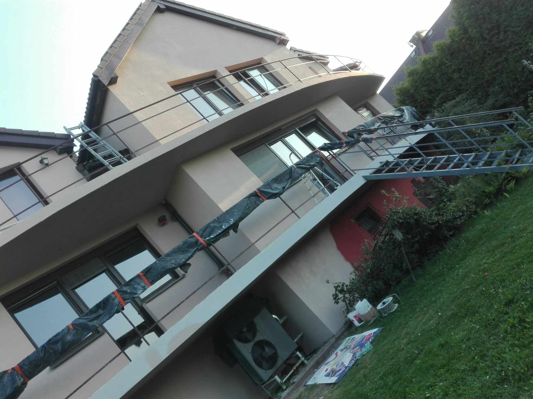 Dwupiętrowy dom z balkonami w trakcie prac elewacyjnych, widoczna drabina zabezpieczona folią, częściowo pomalowana ściana i sprzęt na trawniku.