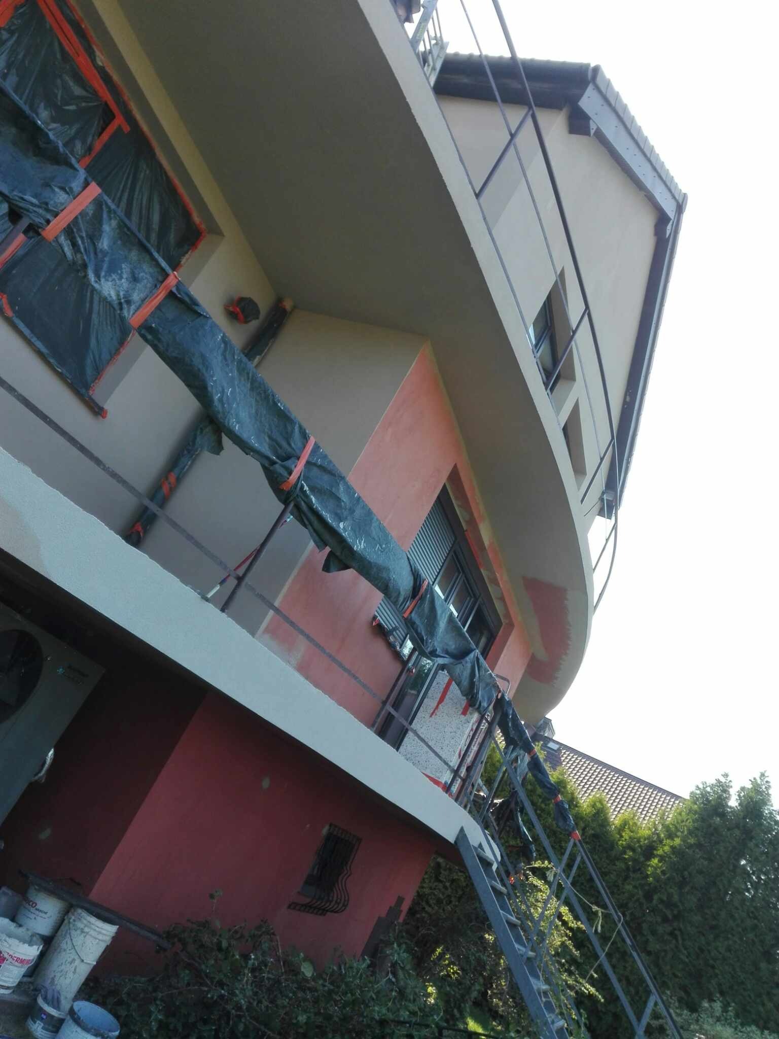 Budynek z częściowo ocieploną elewacją, okna zabezpieczone folią, widoczna drabina i wiaderka z farbą, budynek w trakcie prac remontowych.