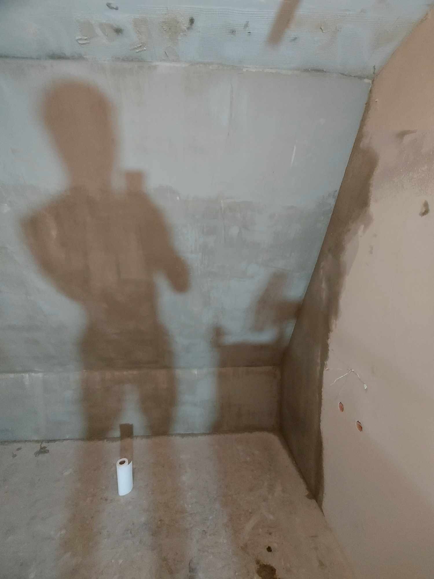 Ściana pod skosem w trakcie prac wykończeniowych, widoczna siatka na suficie, ślady po gładzi, cień osoby robiącej zdjęcie i rolka papieru na podłodze.
