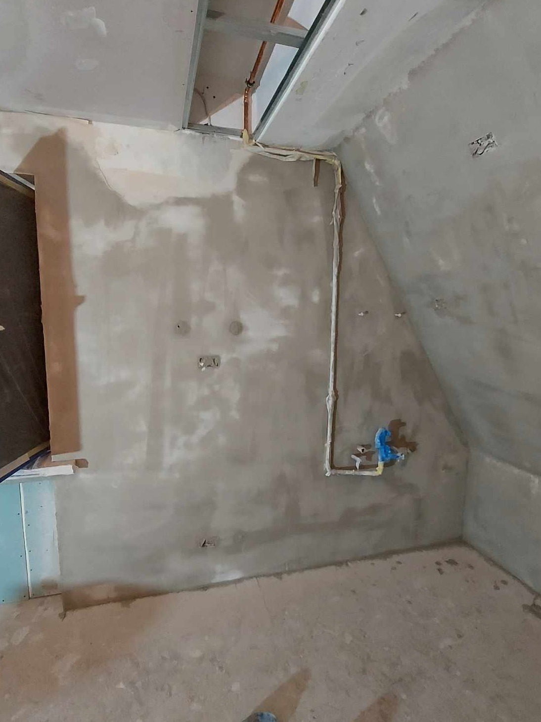 Surowa ściana po nałożeniu gładzi, widoczne rury instalacji wodnej zabezpieczone folią, metalowa konstrukcja sufitu podwieszanego i fragment ściany z płyt gipsowo-kartonowych.
