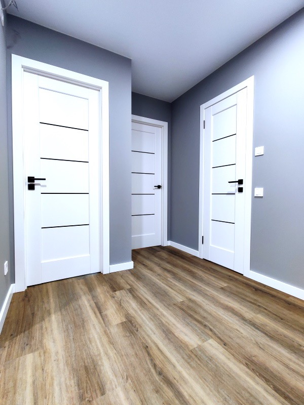 Wnętrze z trzema białymi drzwiami z czarnymi poziomymi paskami i klamkami, szare ściany i drewniana podłoga ułożona wzdłuż korytarza.