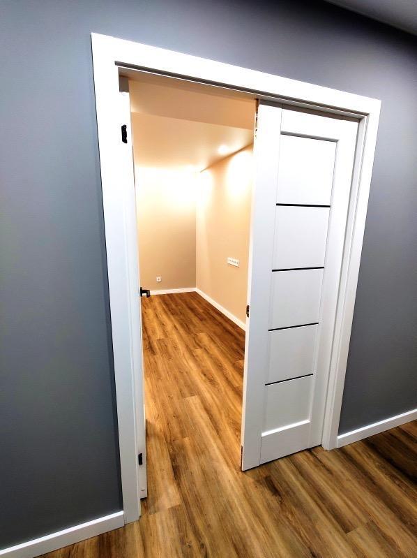 Wnętrze pokoju z drewnianą podłogą, widok przez przesuwne białe drzwi z czarnymi akcentami, ściany pomalowane na szaro i beżowo.