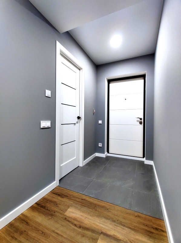 Wnętrze przedpokoju w szarościach z dwiema białymi drzwiami o minimalistycznym designie, widoczne gniazdka elektryczne i kontrastowe podłogi: drewniana i z szarych płytek.