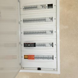 NRG Norbert Radosz - Otwarta skrzynka rozdzielcza z bezpiecznikami i wyłącznikami różnicowoprądowymi w białej obudowie, zamontowana na beżowej ścianie. Widoczne rzędy bezpieczników marki Eaton i Adelid oraz przełącznik...