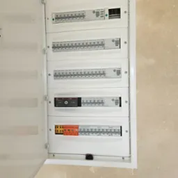 Otwarta skrzynka rozdzielcza z bezpiecznikami i wyłącznikami r&oacute;żnicowoprądowymi w białej obudowie, zamontowana na beżowej ścianie. Widoczne rzędy bezpiecznik&oacute;w marki Eaton i Adelid oraz przełącznik...