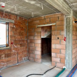 NRG Norbert Radosz - Surowy stan budynku z widoczną instalacją elektryczną: kable prowadzone po suficie, puszki elektryczne w ścianach z cegły, rury instalacyjne na podłodze.