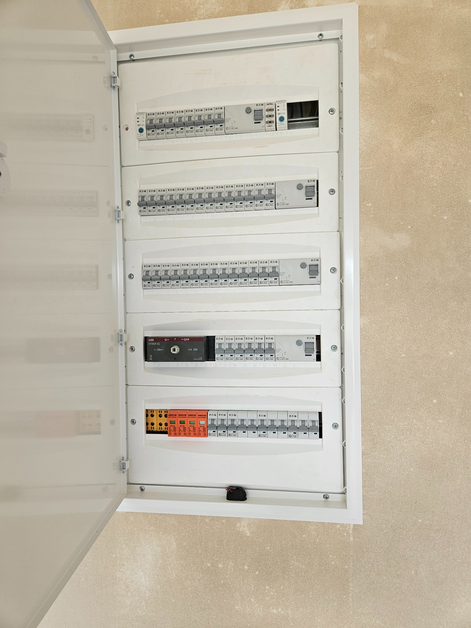 Otwarta skrzynka rozdzielcza z bezpiecznikami i wyłącznikami różnicowoprądowymi w białej obudowie, zamontowana na beżowej ścianie. Widoczne rzędy bezpieczników marki Eaton i Adelid oraz przełącznik...