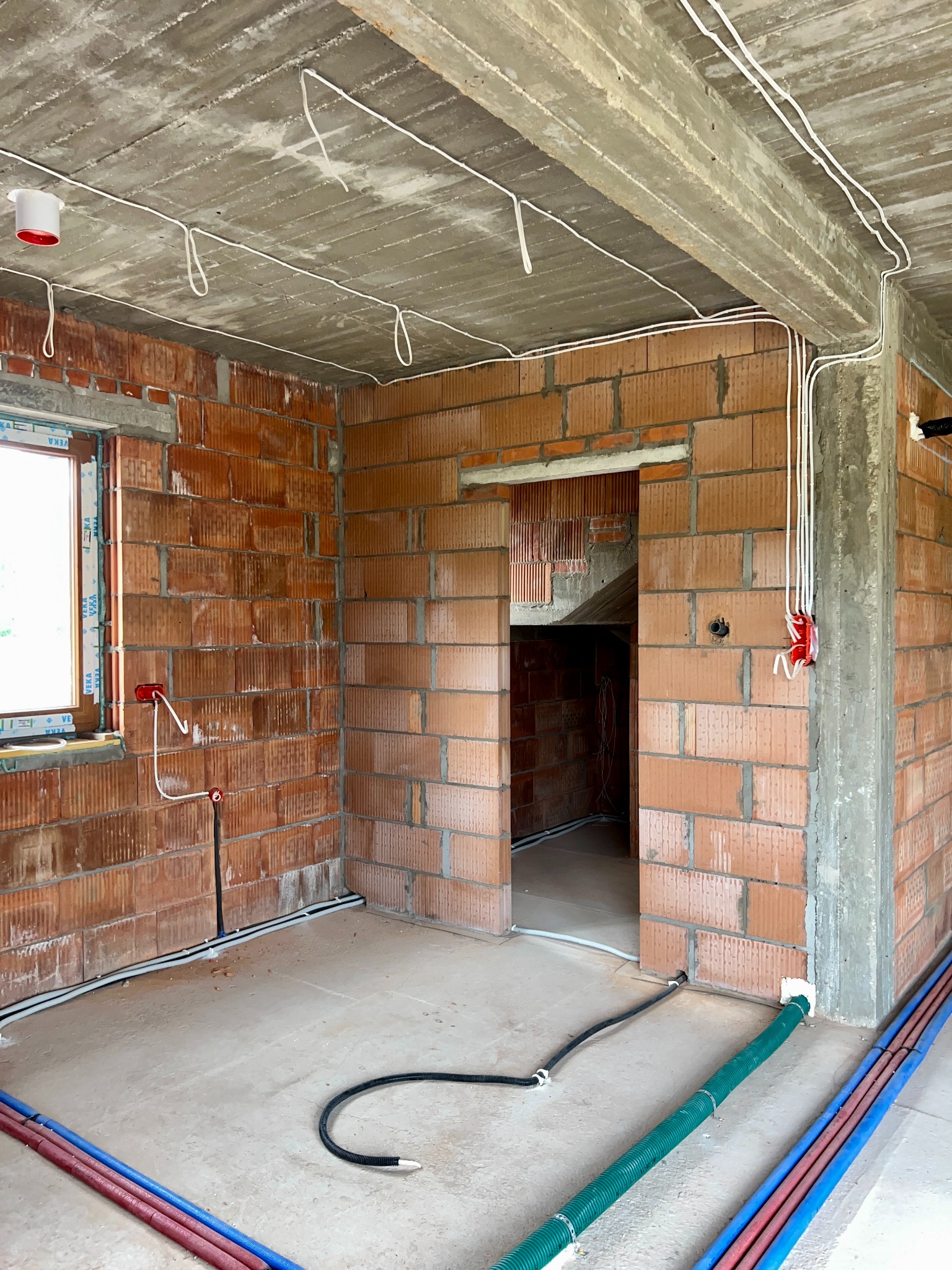 Surowy stan budynku z widoczną instalacją elektryczną: kable prowadzone po suficie, puszki elektryczne w ścianach z cegły, rury instalacyjne na podłodze.