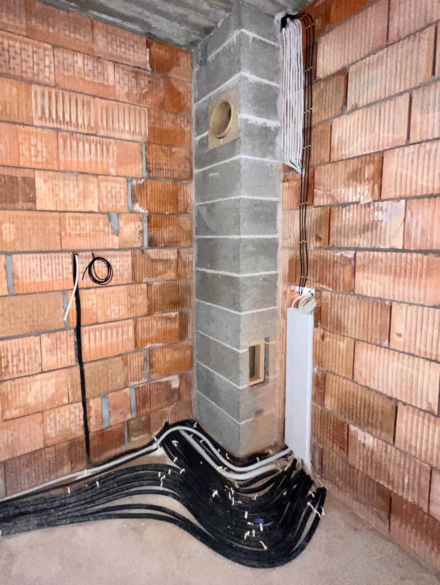 Wewnętrzna instalacja elektryczna w surowym stanie: kable w peszlach ułożone przy ceglanej ścianie i betonowym kominie z otworem wentylacyjnym, przewody wyprowadzone na ścianę w białej listwie.