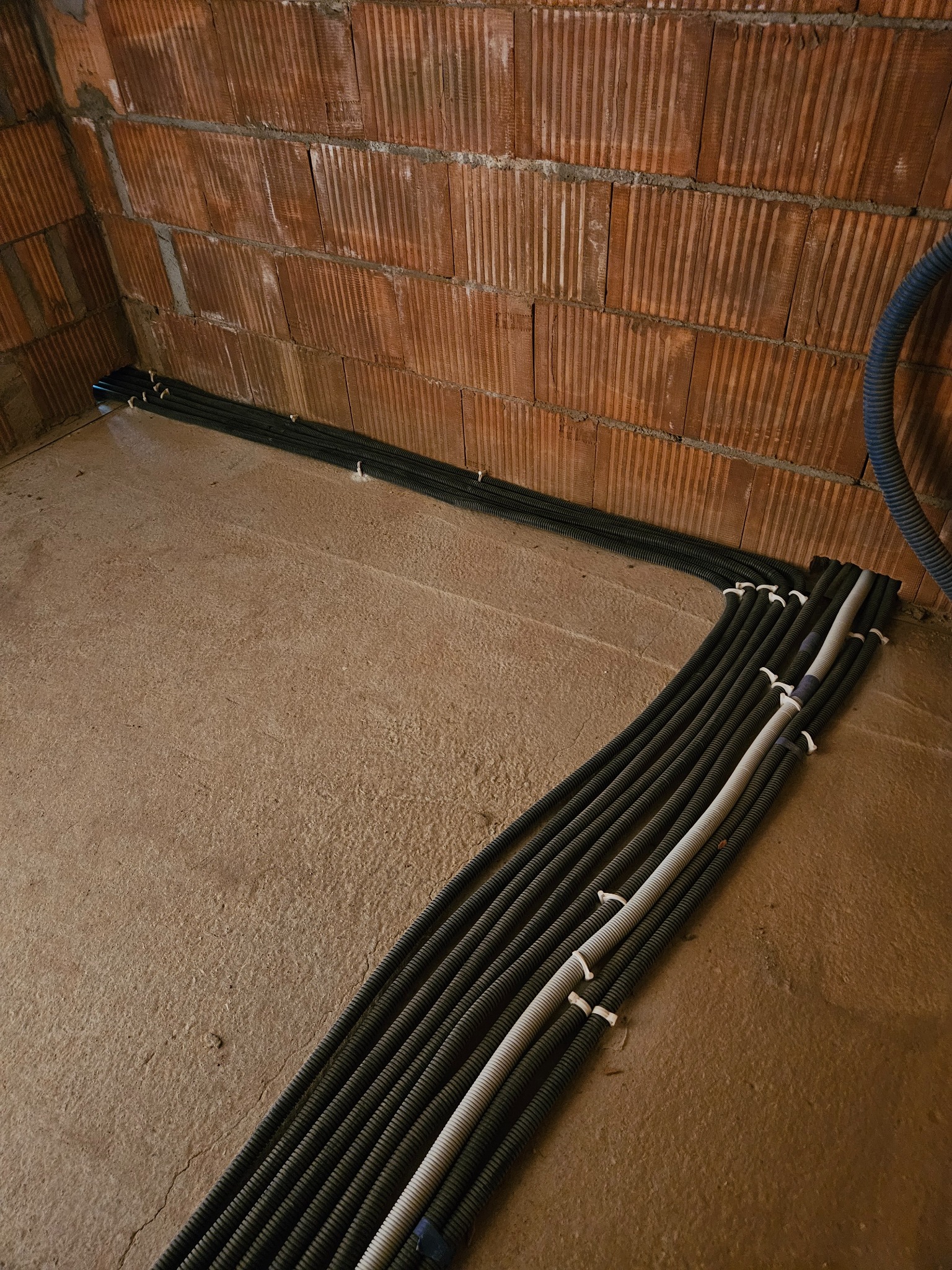 Rzędy czarnych karbowanych peszli z kablami na surowym betonowym podłożu wzdłuż ceglanej ściany, zabezpieczone białymi opaskami zaciskowymi.