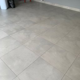 Wojciech Wałek - Jasnoszara podłoga wyłożona kwadratowymi płytkami, widoczne rury instalacyjne pomalowane na czerwono oraz biały zmiękczacz wody i worek na śmieci.